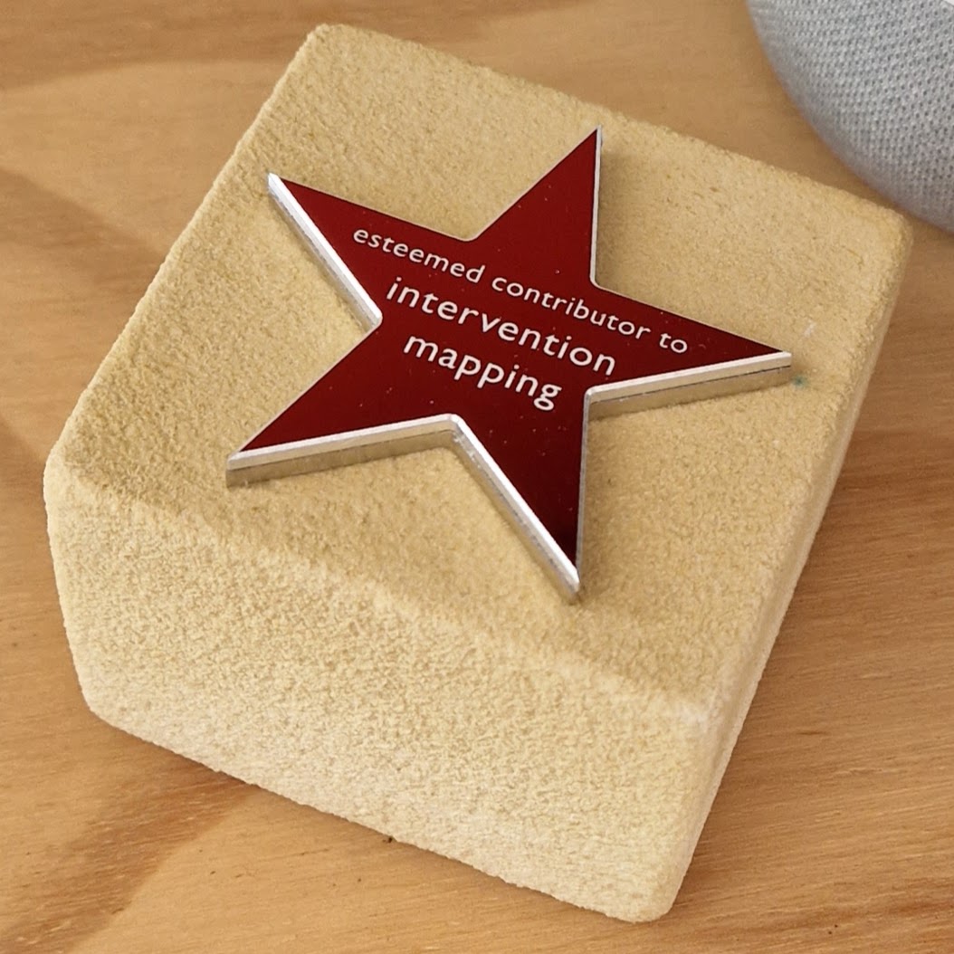 A photograph of a merle stone cube with an aluminum red star on it with the text 'esteemed contributor to intervention mapping' on it.